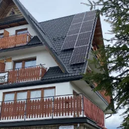 Dwukondygnacyjny dom z kamienną elewacją i drewnianymi balkonami, na dachu zamontowane panele słoneczne, widok z dołu, częściowo zasłonięte przez gałęzie drzewa.