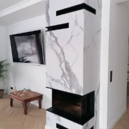 Nowoczesny kominek obudowany płytami imitującymi marmur z czarnymi akcentami, w tle telewizor i parkiet w jodełkę.