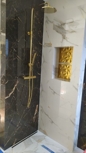 Nowoczesna kabina prysznicowa z czarnymi marmurowymi płytkami i złotą armaturą, akcentowana wnęką z trójwymiarową, złotą teksturą na białej marmurowej ścianie.