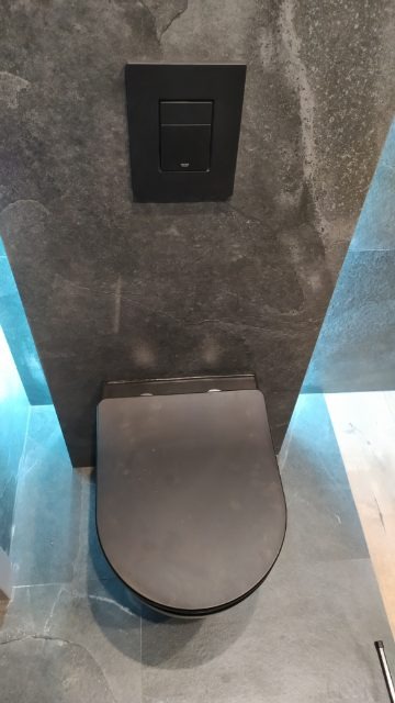 Wisząca, czarna toaleta z czarnym przyciskiem spłukującym na szarej, kamiennej ścianie w nowoczesnej łazience.