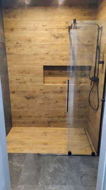 Wykończona kabina prysznicowa z drewnopodobnymi płytkami na ścianach i podłodze, czarna armatura, szklane drzwi z czarnym uchwytem, wnęka w ścianie wykończona tym samym materiałem, szare płytki...