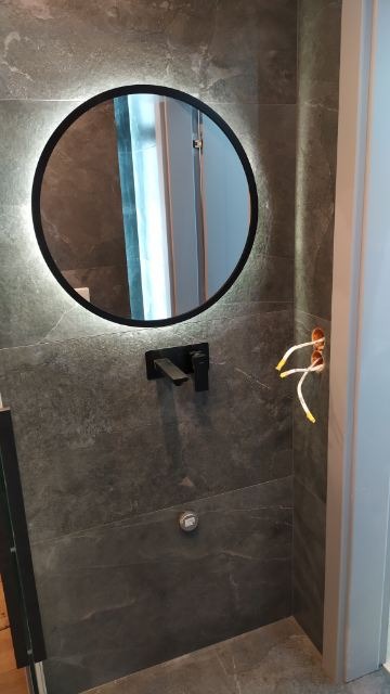 Częściowo wykończona łazienka z okrągłym lustrem podświetlanym LED, czarną armaturą i widocznymi przewodami elektrycznymi w ścianie z szarej płytki.