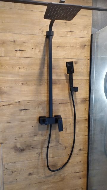 Czarny zestaw natryskowy z deszczownicą i słuchawką, zamontowany na ścianie wyłożonej panelami imitującymi drewno, z fragmentem szarej płyty po prawej stronie.