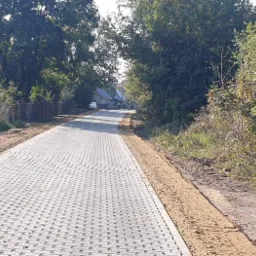 Ułożona szara kostka ażurowa na nowej drodze osiedlowej, z przygotowanym obramowaniem z piasku, widok w perspektywie w kierunku zabudowań.