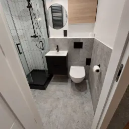 Nowoczesna łazienka z kabiną prysznicową typu walk-in z czarnymi ramami, umywalką z szafką w kolorze czarnym, wiszącą toaletą i szarą glazurą na ścianach.