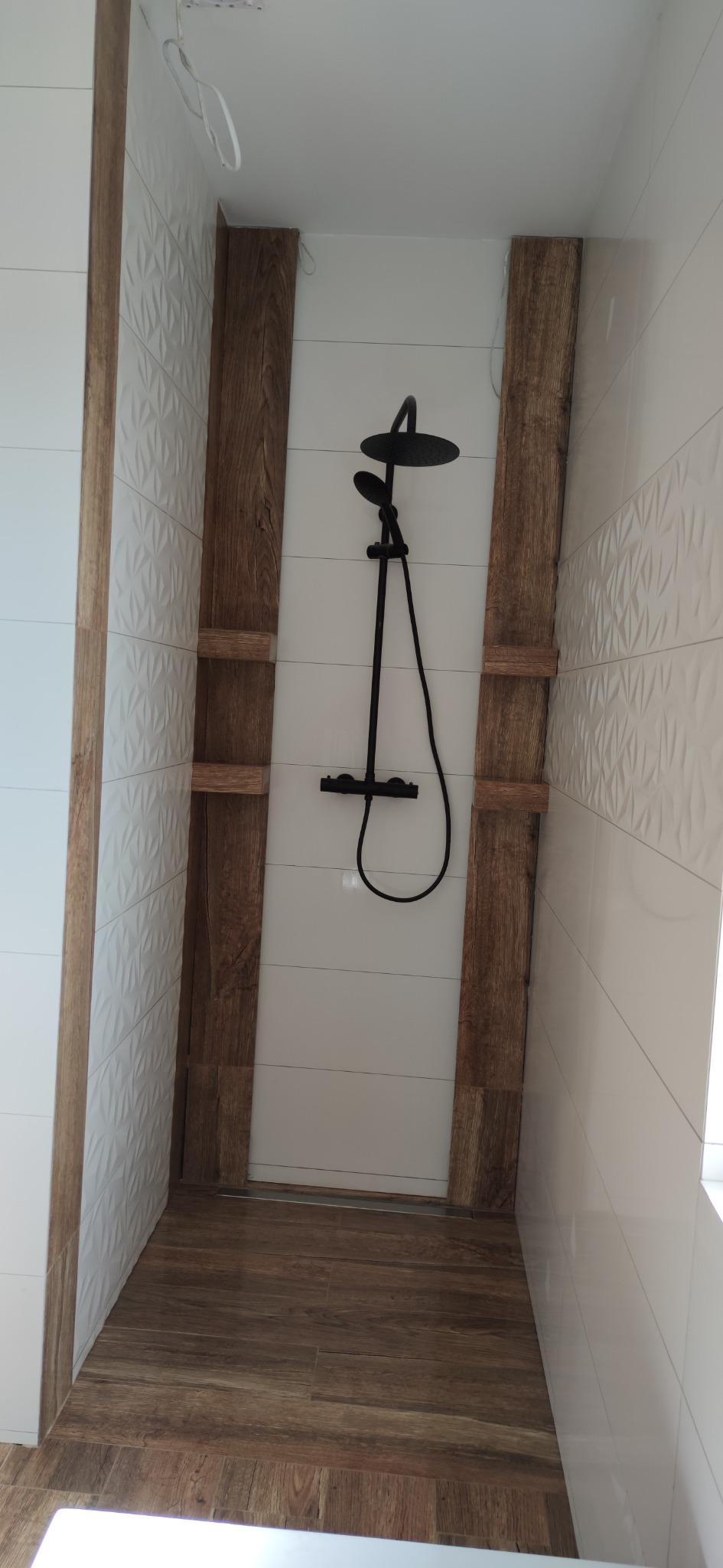 Kabina prysznicowa walk-in z czarną armaturą, obłożona białymi płytkami z trójwymiarowym wzorem i drewnopodobnymi panelami oraz podłogą, widok z góry.