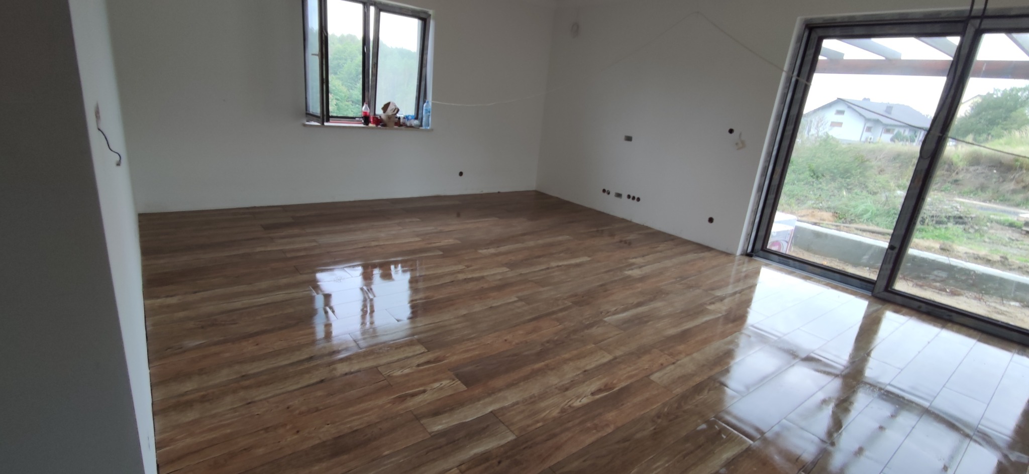 Salon z nową, błyszczącą podłogą imitującą drewno, duże okno przesuwne z widokiem na zewnątrz, ściany w trakcie wykończenia, widoczne puszki elektryczne.