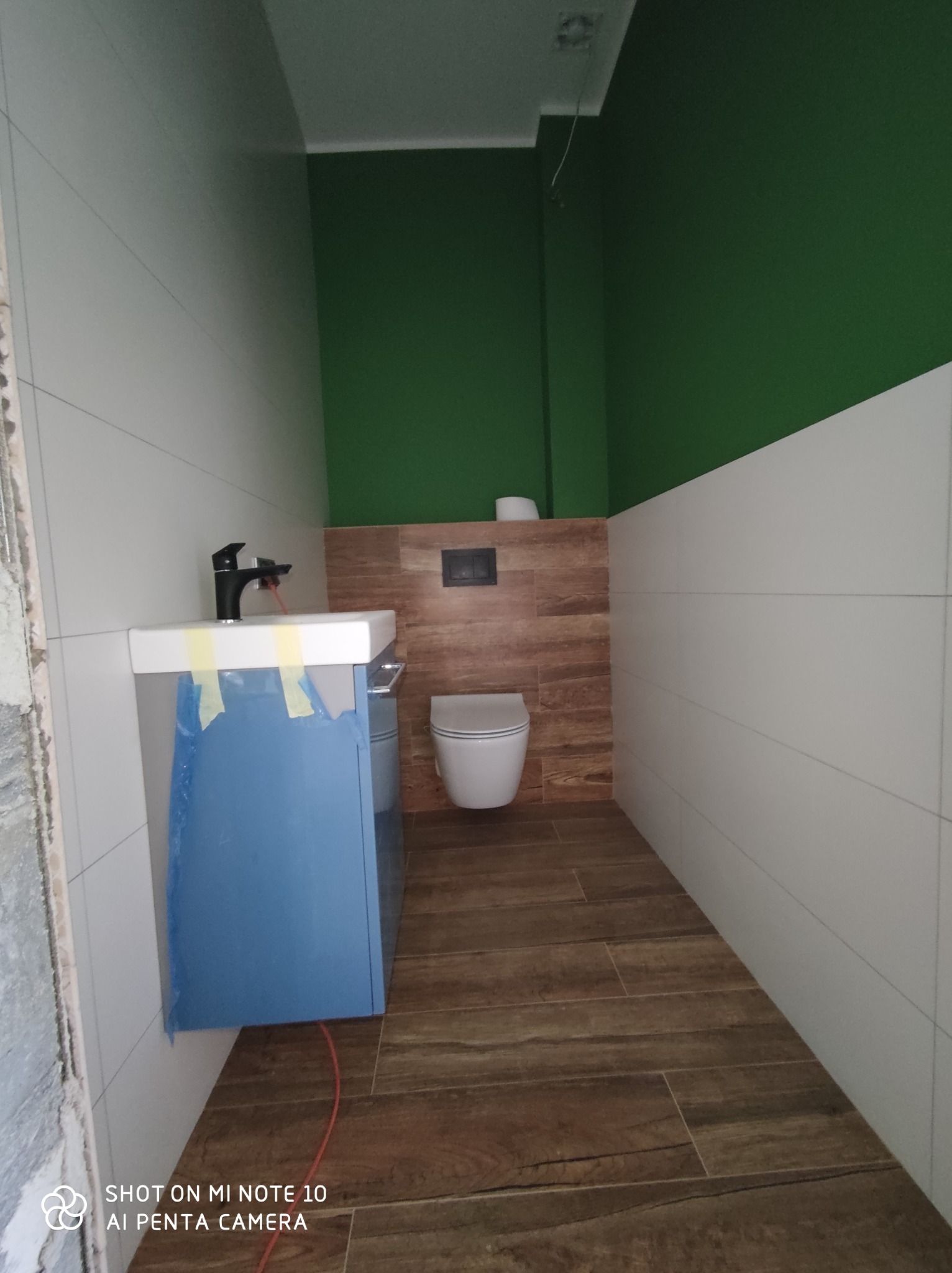 Łazienka w trakcie wykańczania: białe płytki ścienne, drewnopodobna podłoga, toaleta wisząca, zielona ściana w górnej części, umywalka z szafką zabezpieczona folią ochronną.