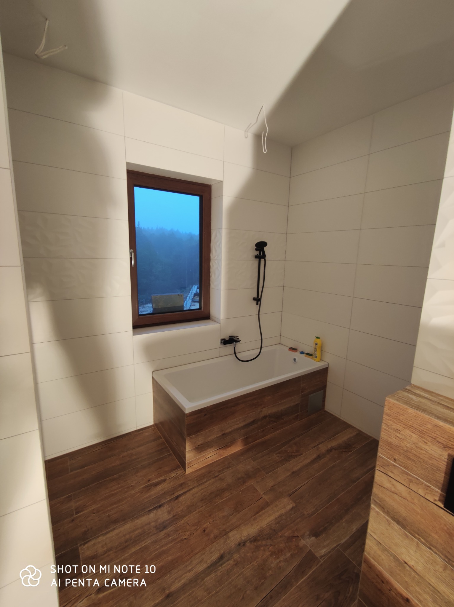 Nowoczesna łazienka w Jastrzębiu Zdroju z białymi płytkami imitującymi fale, drewnopodobną podłogą i obudową wanny, oraz oknem z widokiem na zalesione wzgórze.