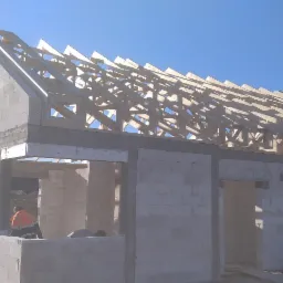 Budowa domu murowanego z pustaków: wzniesione ściany i konstrukcja dachu z drewna, widoczni pracownicy wewnątrz budynku, na szczycie dachu wieniec, jasne niebo w tle.