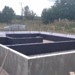 Zalane fundamenty domu z widoczną izolacją pionową, zbrojeniami i szalunkiem traconym w otoczeniu zieleni i drzew.