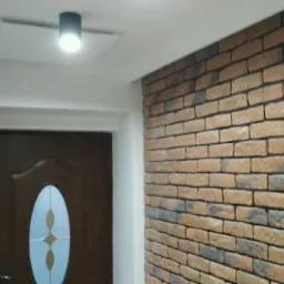 Ściana wykończona cegłą dekoracyjną w odcieniach beżu i brązu, przylegająca do białej ściany z drzwiami wejściowymi z brązowym wzorem i owalnym przeszkleniem, oświetlona nowoczesną lampą sufitową.