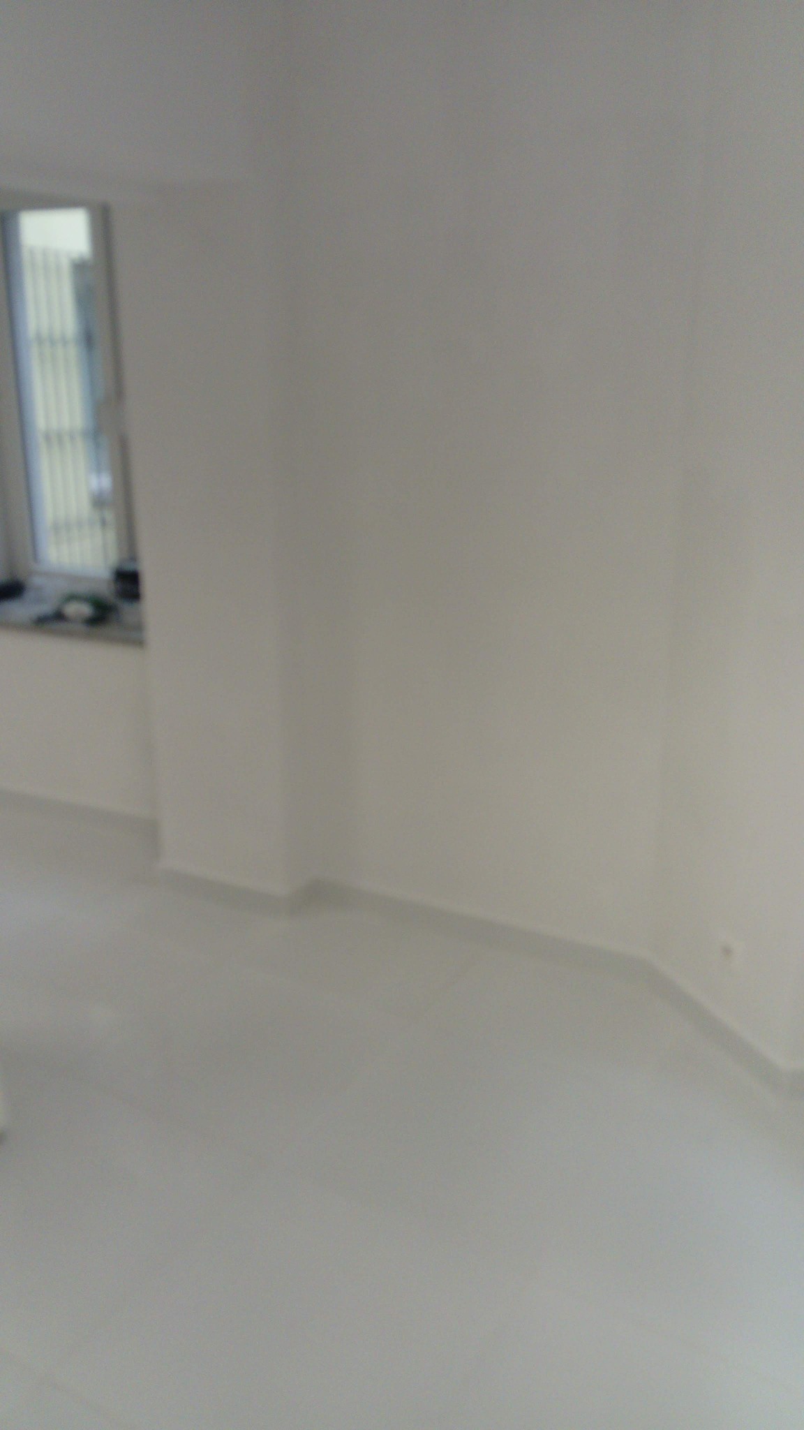 Świeżo wyremontowane, jasne pomieszczenie z białymi ścianami, podłogą i listwami przypodłogowymi. Okno z żaluzjami w lewym górnym rogu. Rozmycie sugeruje niedawne prace wykończeniowe lub sprzątanie.