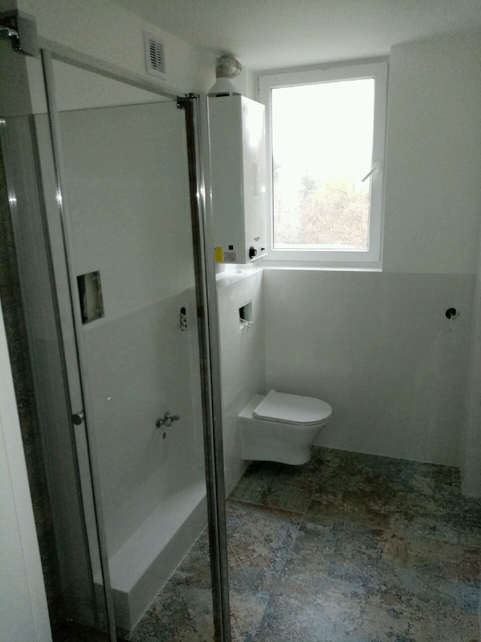 Nowoczesna łazienka z kabiną prysznicową ze szklanymi drzwiami, podwieszaną toaletą i piecem gazowym wiszącym nad nią, wykończona płytkami imitującymi wzorzysty dywan.