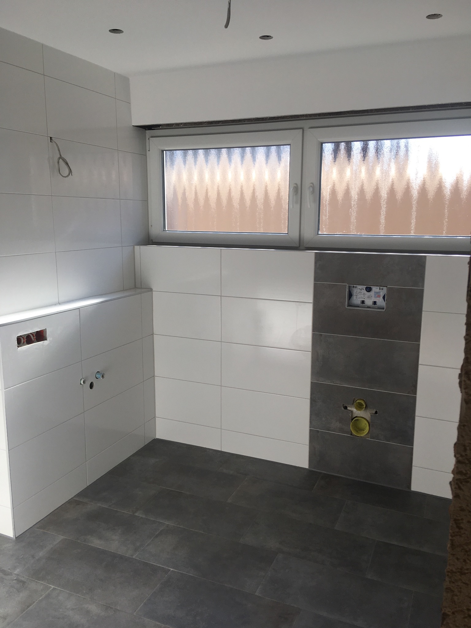 Wnętrze łazienki w trakcie remontu, widoczne białe i szare płytki na ścianach, szare płytki podłogowe, okno z szybą ornamentową oraz instalacja sanitarna w ścianie.