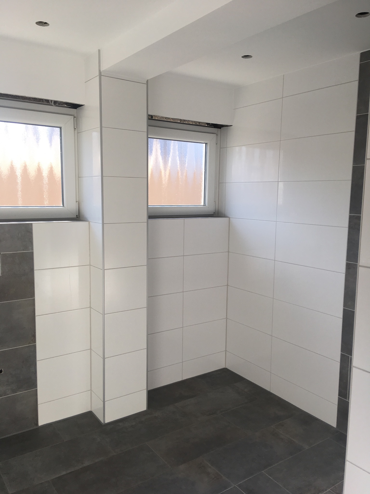 Wykończona łazienka z białymi płytkami na ścianach i ciemnoszarą podłogą, widoczne dwa okna z białymi ramami i fragment listwy dekoracyjnej w kolorze szarym.