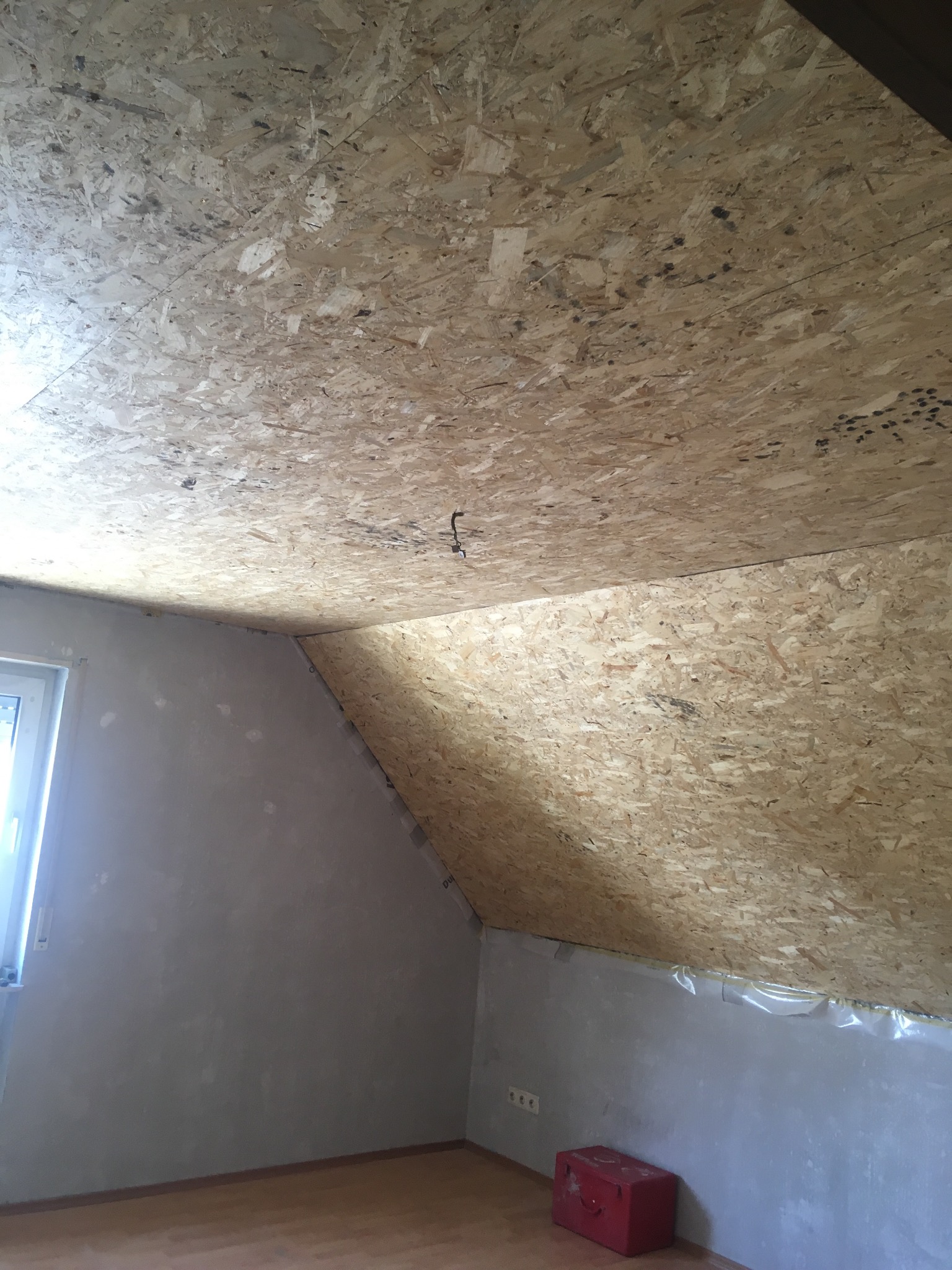 Poddasze w trakcie prac wykończeniowych: sufit pokryty płytami OSB, ściany przygotowane do malowania, widoczna instalacja elektryczna i czerwona skrzynka narzędziowa przy ścianie.