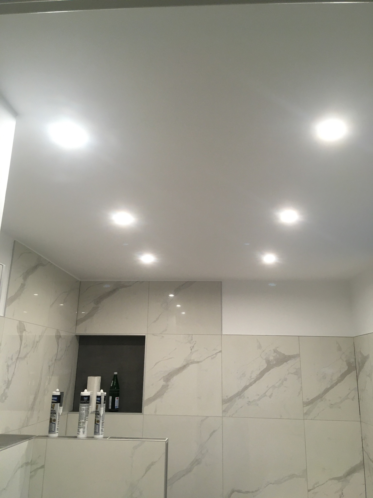 Wykończona łazienka z jasnymi płytkami imitującymi marmur, wbudowana półka z butelką i akcesoriami montażowymi, oświetlenie sufitowe punktowe.