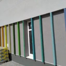 Ocieplona elewacja budynku z kolorowymi, pionowymi pasami dekoracyjnymi, paleta worków z zaprawą Weber na pierwszym planie.