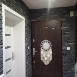 Wnętrze przedpokoju z dekoracyjną ścianą wyłożoną szarym kamieniem, widoczne białe drzwi wewnętrzne i ciemne drzwi wejściowe z zawieszonym łapaczem sn&oacute;w.