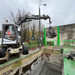 KORMOL Grzegorz Korda - Koparka Mecalac z łyżką zawieszoną nad głębokim wykopem o nierównych ścianach, zabezpieczonym zielonymi barierami, operator w kamizelce odblaskowej, tło z drzewami i słupem oświetleniowym.