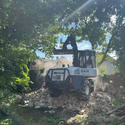 KORMOL Grzegorz Korda - Biała koparka Mecalac stoi na stercie gruzu ceglanego, z tyłu widoczny fragment rozbieranej ceglanej ściany, w tle zarośla i budynek, promienie słoneczne przebijają przez korony drzew.