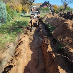 KORMOL Grzegorz Korda - Wąski wykop w ziemi, zabezpieczony drewnianymi stemplami, wykonywany przez koparkę Mecalac w słoneczny dzień na działce z widoczną zielenią i drzewami w tle.