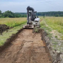 Koparka kołowa w trakcie wykonywania wykopu liniowego w terenie trawiastym, widoczne ślady pracy maszyny na dnie wykopu, w tle drzewa i słupy energetyczne.