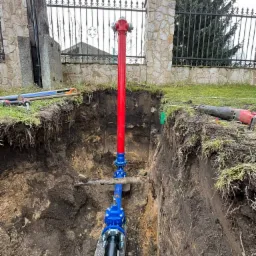 Czerwony hydrant i niebieskie rury wodociągowe zainstalowane w głębokim wykopie ziemnym, widoczne narzędzia budowlane na powierzchni gruntu, w tle ogrodzenie z kamienia i kutego żelaza.