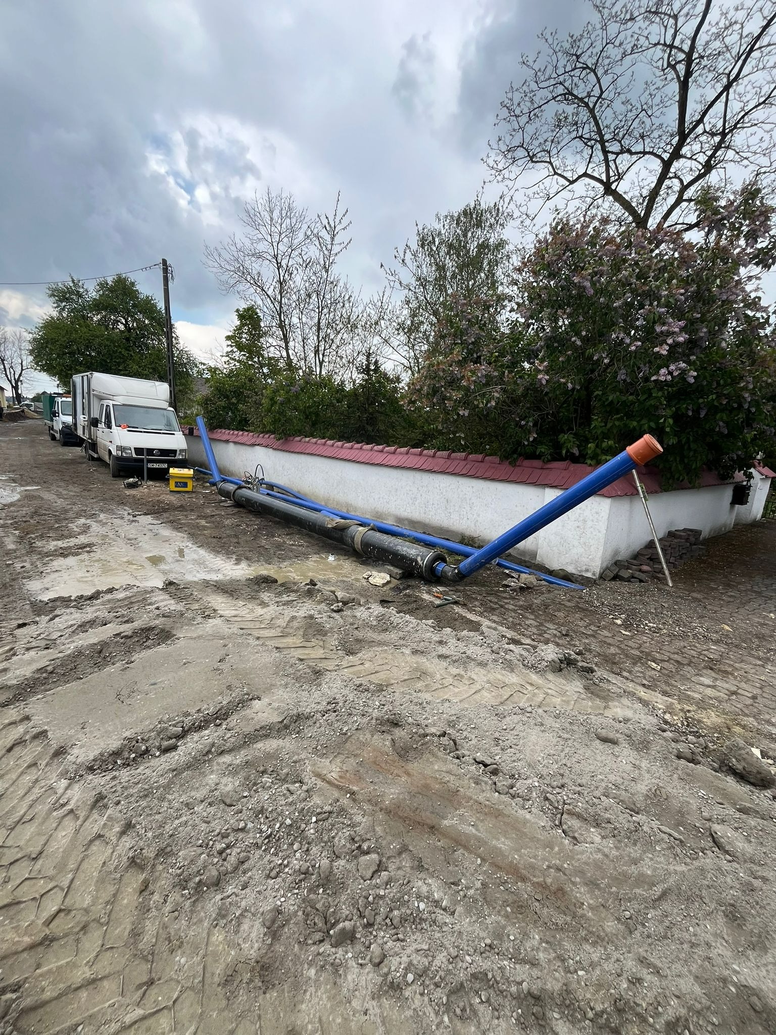 Rury wodociągowe, częściowo niebieskie, leżące na błotnistym terenie obok białego murku z czerwoną dachówką; w tle zaparkowane busy i drzewa pod pochmurnym niebem.