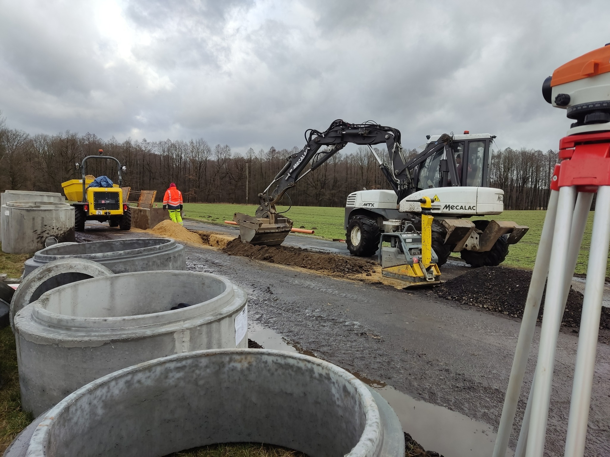 Ciężka koparka Mecalac MTX kopie ziemię na poboczu drogi, obok leżą betonowe kręgi, w tle widoczny żółty ciągnik i pracownik w pomarańczowej kurtce, a w prawym rogu geodezyjny niwelator.