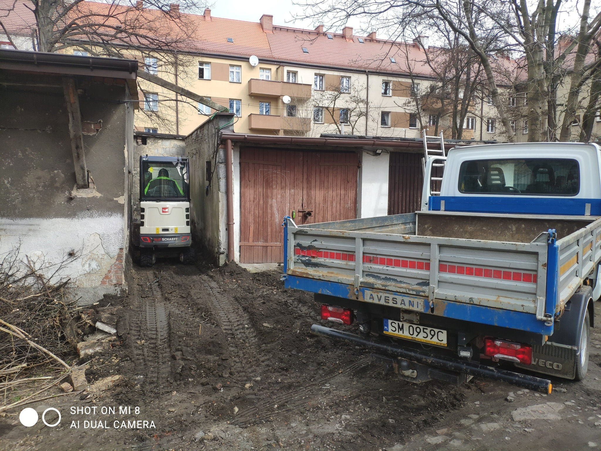 Mini koparka Schaeff w wąskim przejeździe między budynkami, za nią zaparkowany samochód dostawczy Iveco; teren po wykopach, błoto, gałęzie.