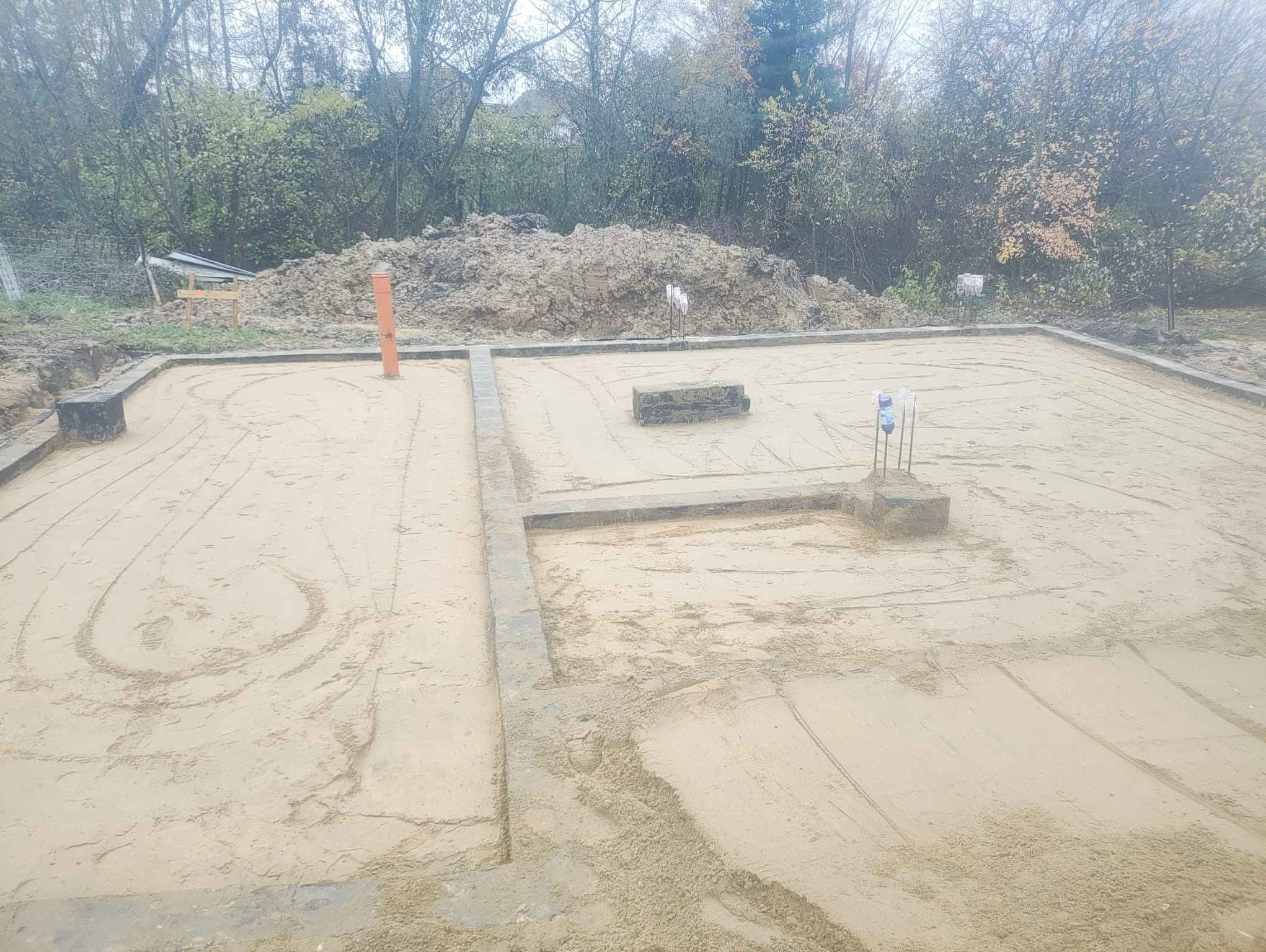 Przygotowany teren pod wylewkę fundamentów z widoczną instalacją kanalizacyjną i sterczącymi prętami zbrojeniowymi, otoczony wykopanym gruntem i drzewami.