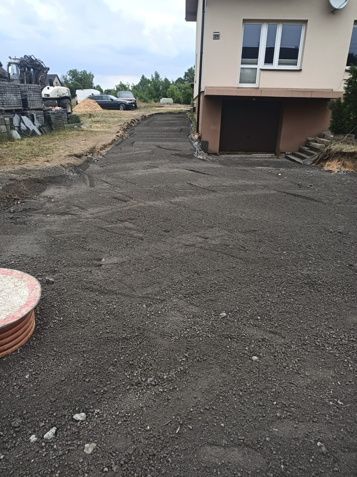 Przygotowany podjazd do domu z wysypanym i wyrównanym tłuczniem, widoczny budynek z garażem w tle, po lewej stronie maszyny budowlane i materiały.