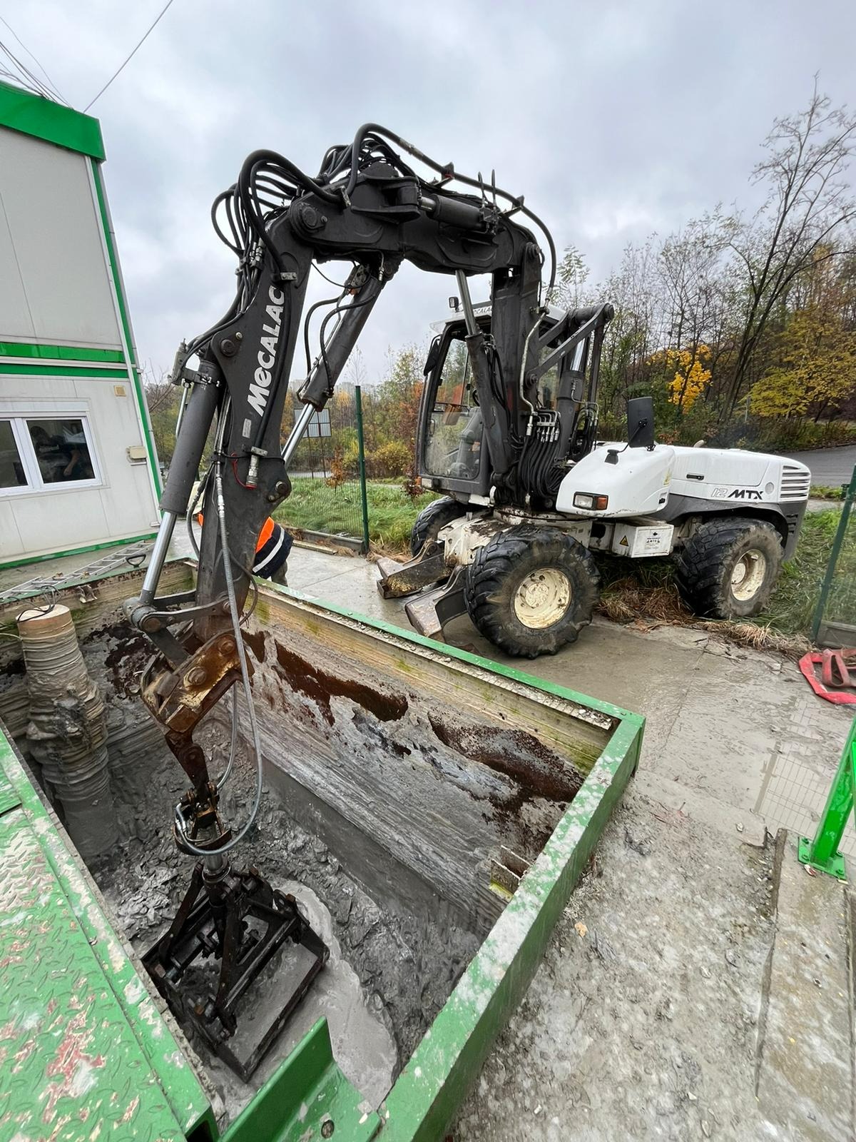 Kołowa koparka Mecalac z chwytakiem w zielonej obudowie wykopu, przygotowująca otwór w ziemi pod instalacje; widoczny budynek w tle