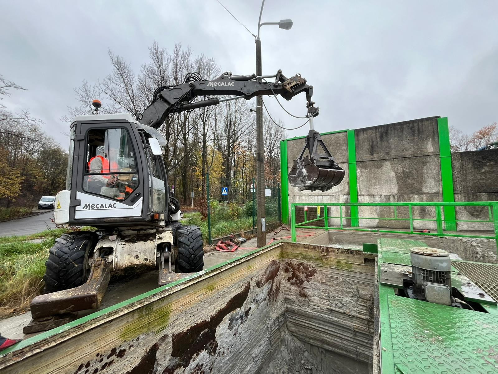 Koparka Mecalac z łyżką zawieszoną nad głębokim wykopem o nierównych ścianach, zabezpieczonym zielonymi barierami, operator w kamizelce odblaskowej, tło z drzewami i słupem oświetleniowym.