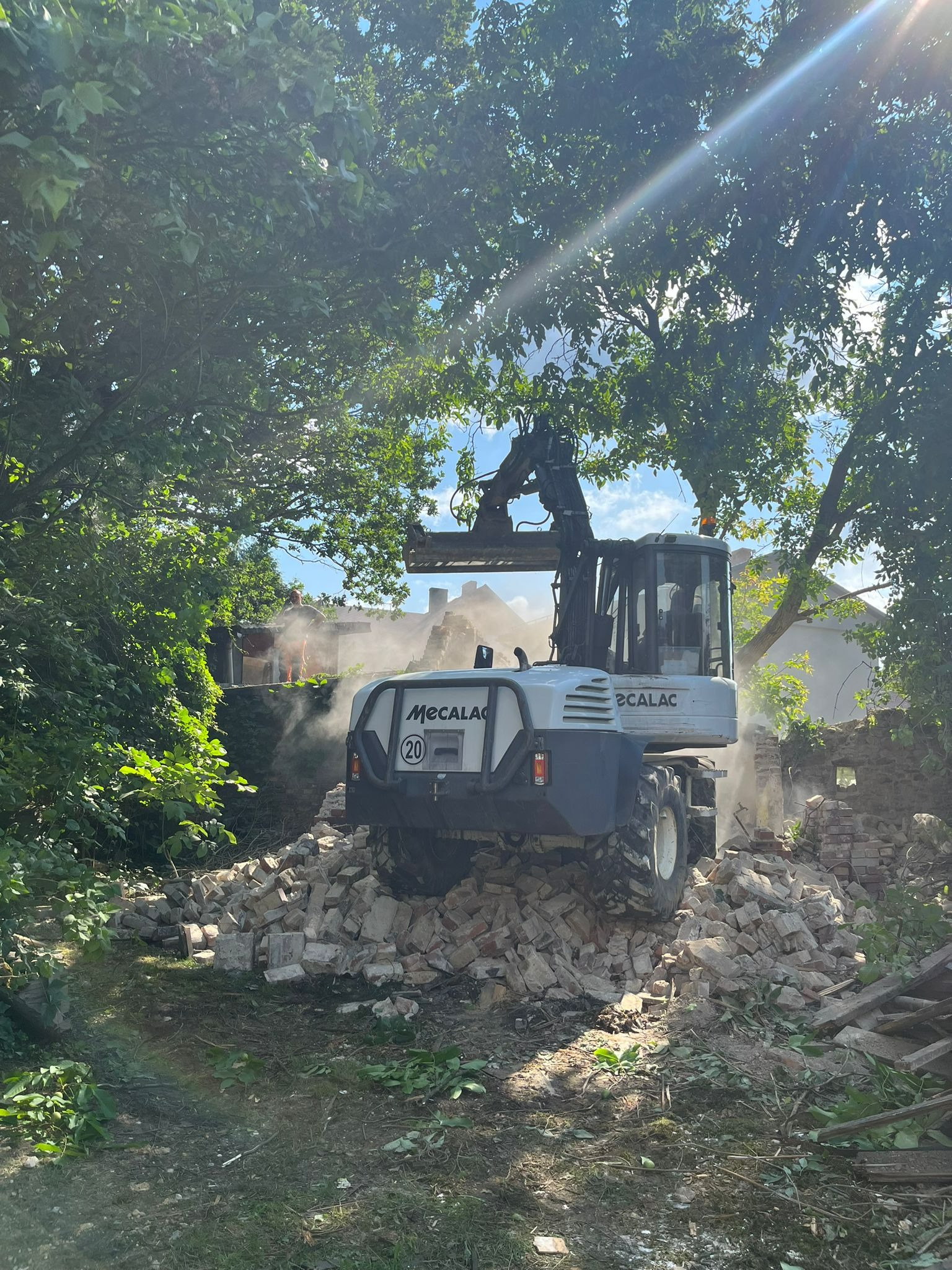 Biała koparka Mecalac stoi na stercie gruzu ceglanego, z tyłu widoczny fragment rozbieranej ceglanej ściany, w tle zarośla i budynek, promienie słoneczne przebijają przez korony drzew.