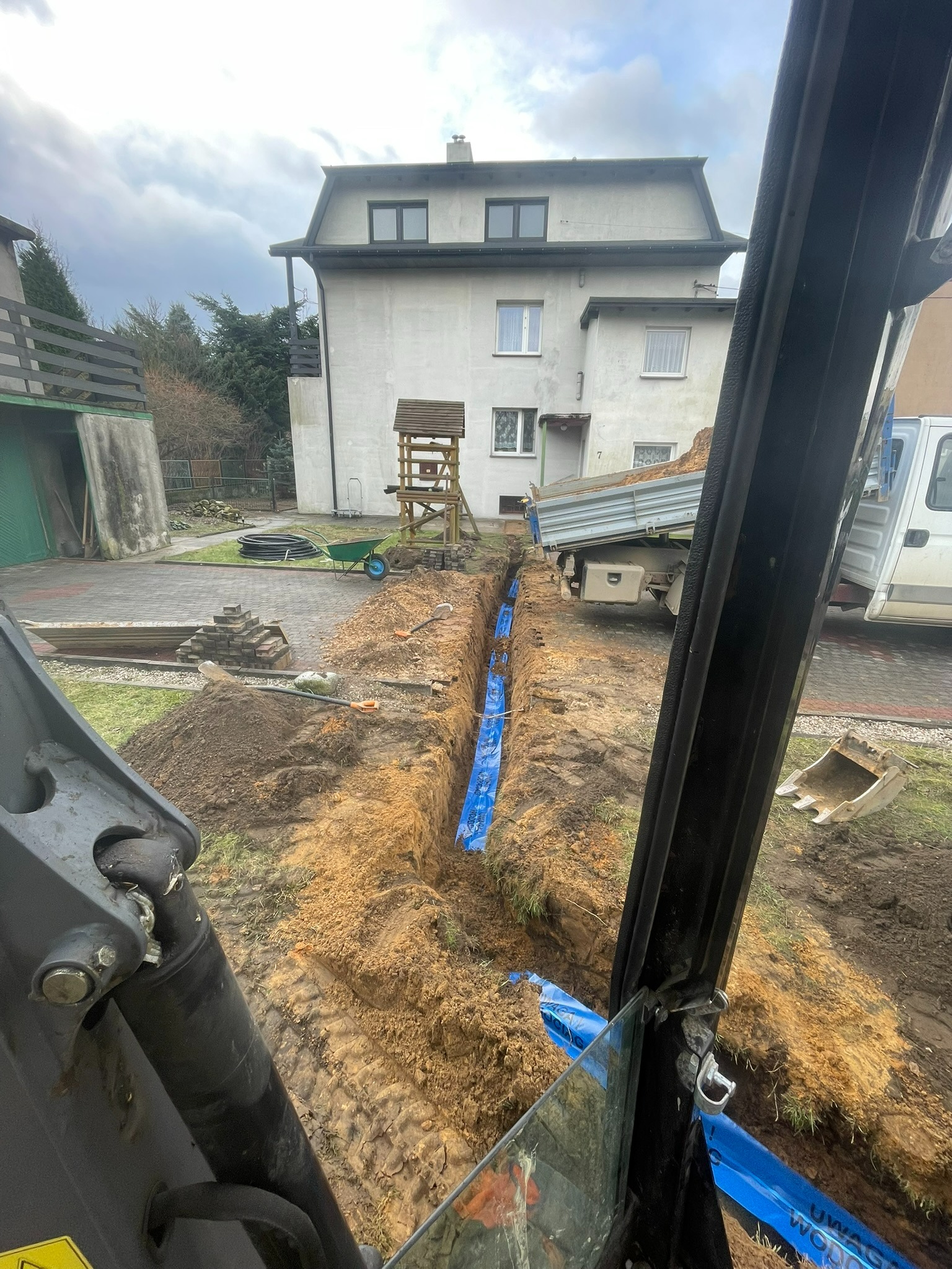 Widok z kabiny koparki na wykop w ziemi z niebieską folią zabezpieczającą, w tle dom jednorodzinny i wywrotka z piaskiem.