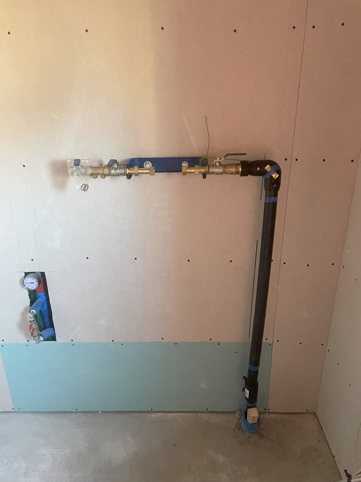 Nowe przyłącze wodno-kanalizacyjne z zaworem, zamontowane na ścianie z płyt gipsowo-kartonowych, z widocznym fragmentem instalacji w ścianie i pionową rurą odpływową do podłogi.