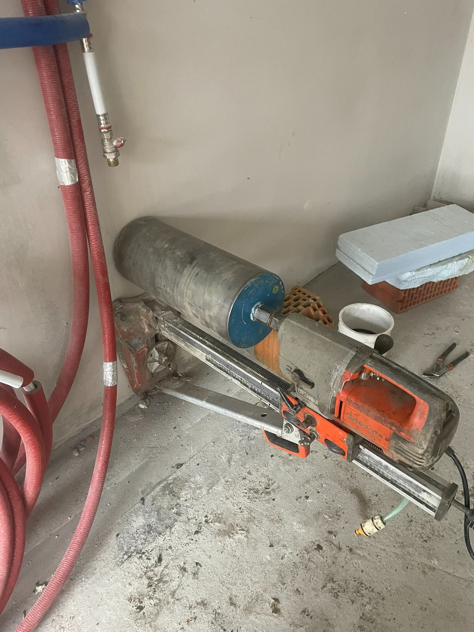 Wiertnica diamentowa Husqvarna z wiertłem koronowym, przygotowana do wiercenia w ścianie, obok widoczne rury instalacji wodnej i materiały budowlane.