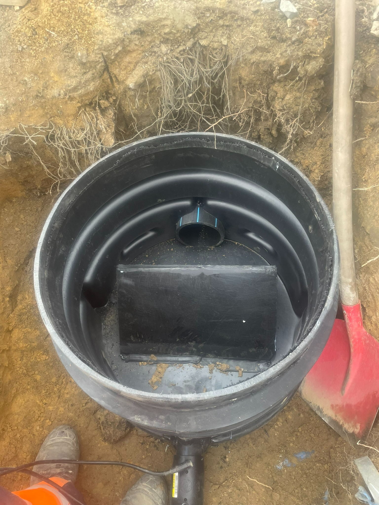 Czarny zbiornik z rurą w wykopie, widoczna łopata i fragment buta osoby nadzorującej, ziemia z korzeniami.