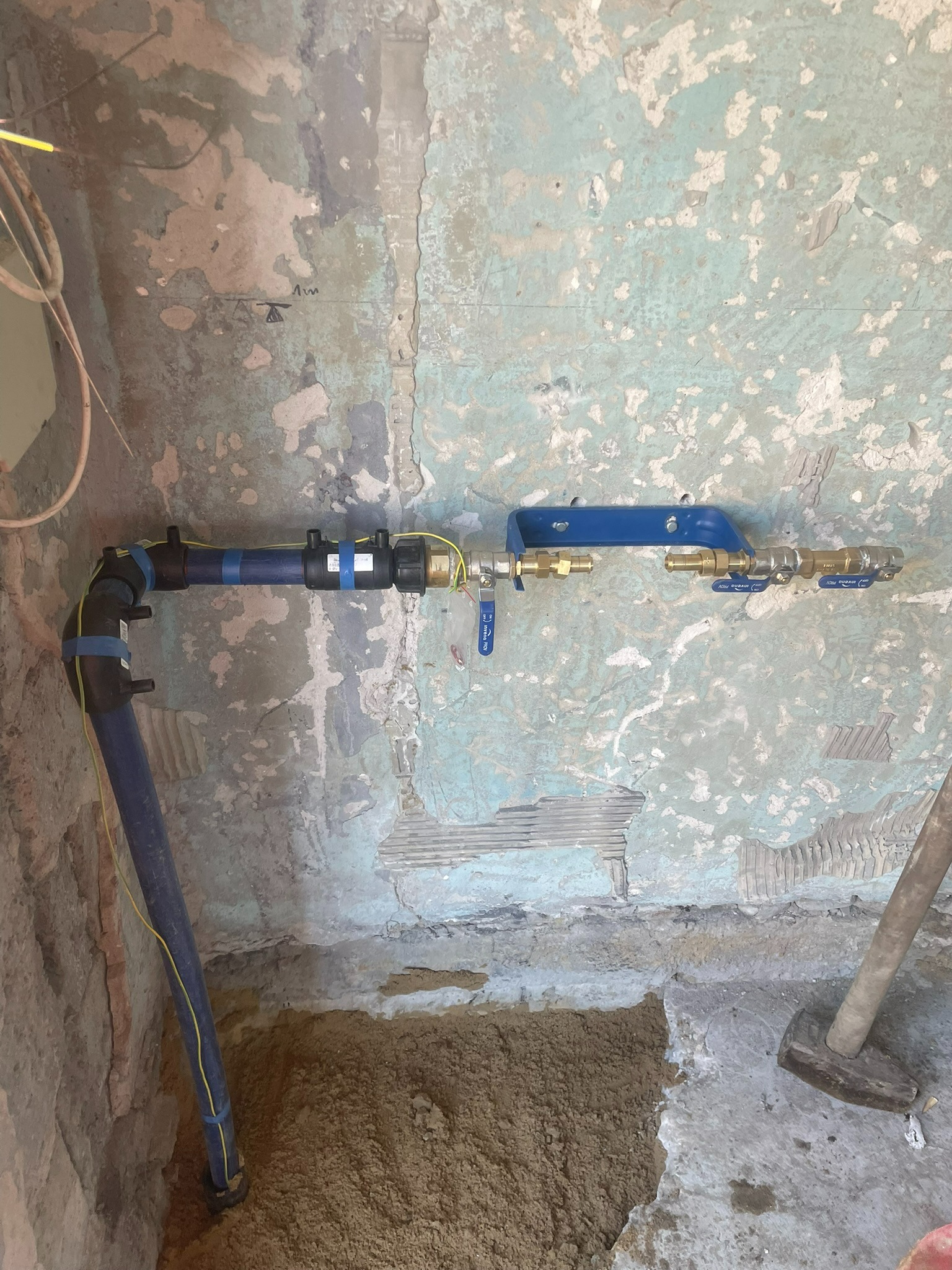 Niebieska rura z podłączeniami wodnymi zamontowana na nierównej, łuszczącej się ścianie, z widocznym wykopem w ziemi i młotem w tle.