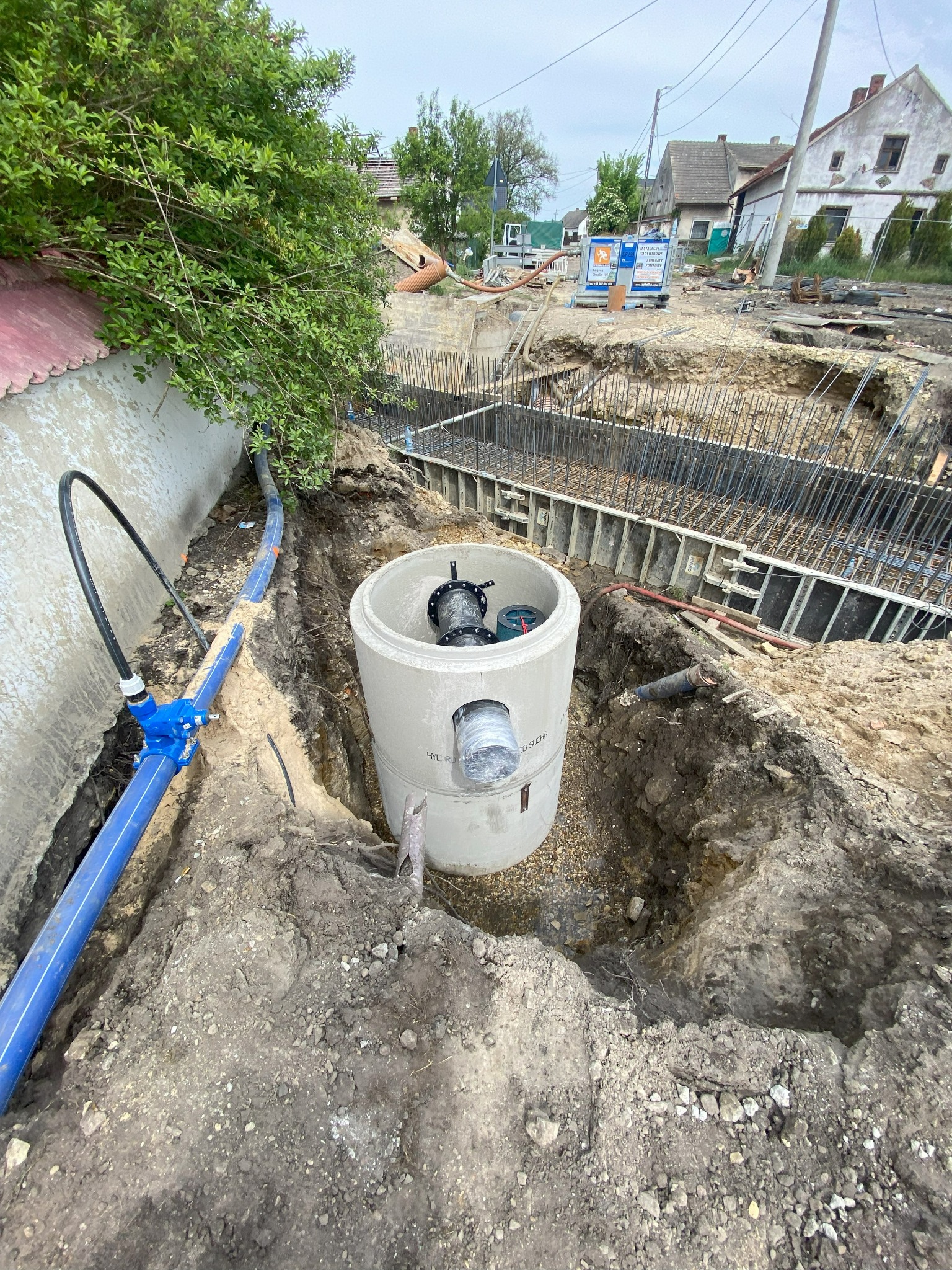 Betonowy krąg studni kanalizacyjnej w wykopie, z widocznymi przyłączami rur i niebieskim rurociągiem wodnym, w tle fundamenty budynku w trakcie budowy.