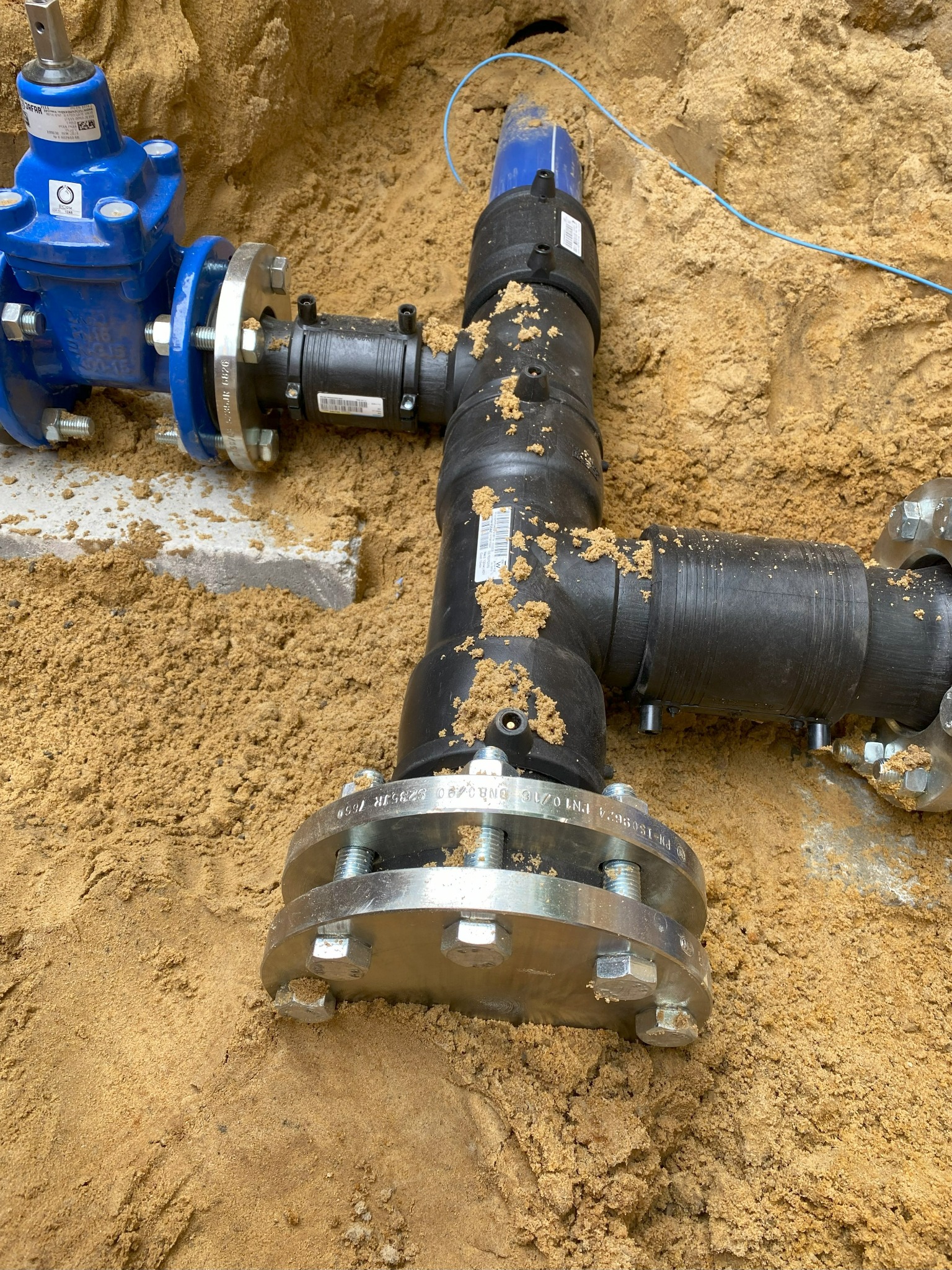 Połączenie rur z kołnierzami i zaworem w wykopie zasypanym piaskiem. Widoczne ślady piasku na elementach instalacji.