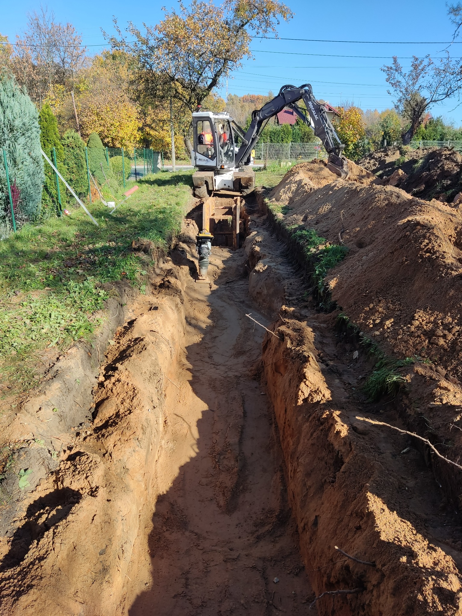 Wąski wykop w ziemi, zabezpieczony drewnianymi stemplami, wykonywany przez koparkę Mecalac w słoneczny dzień na działce z widoczną zielenią i drzewami w tle.