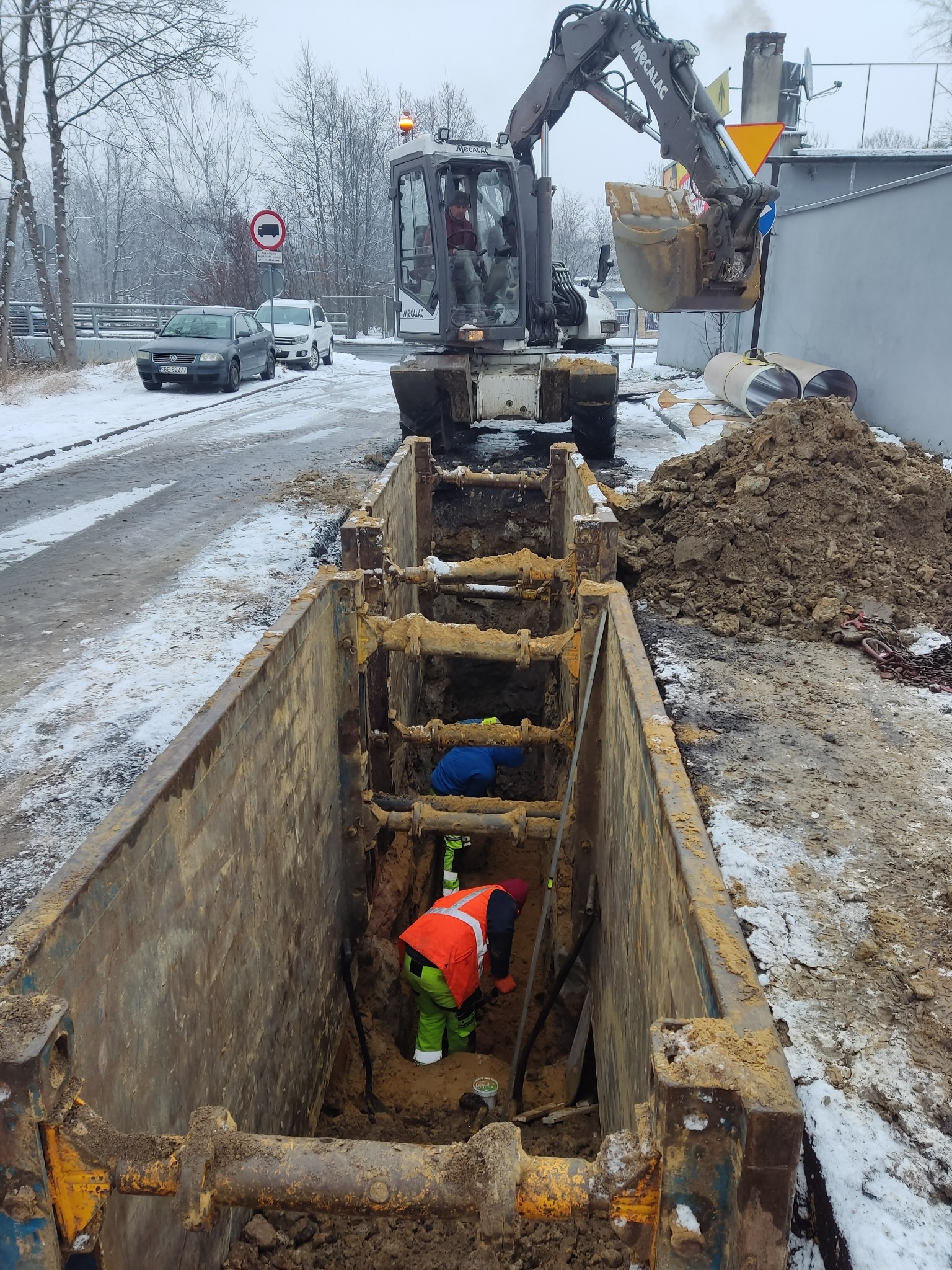 Głęboki wykop zabezpieczony metalowymi szalunkami, robotnicy w odblaskowych ubraniach pracują wewnątrz, koparka MecaLac obok, rury na górze, zimowa sceneria z resztkami śniegu.