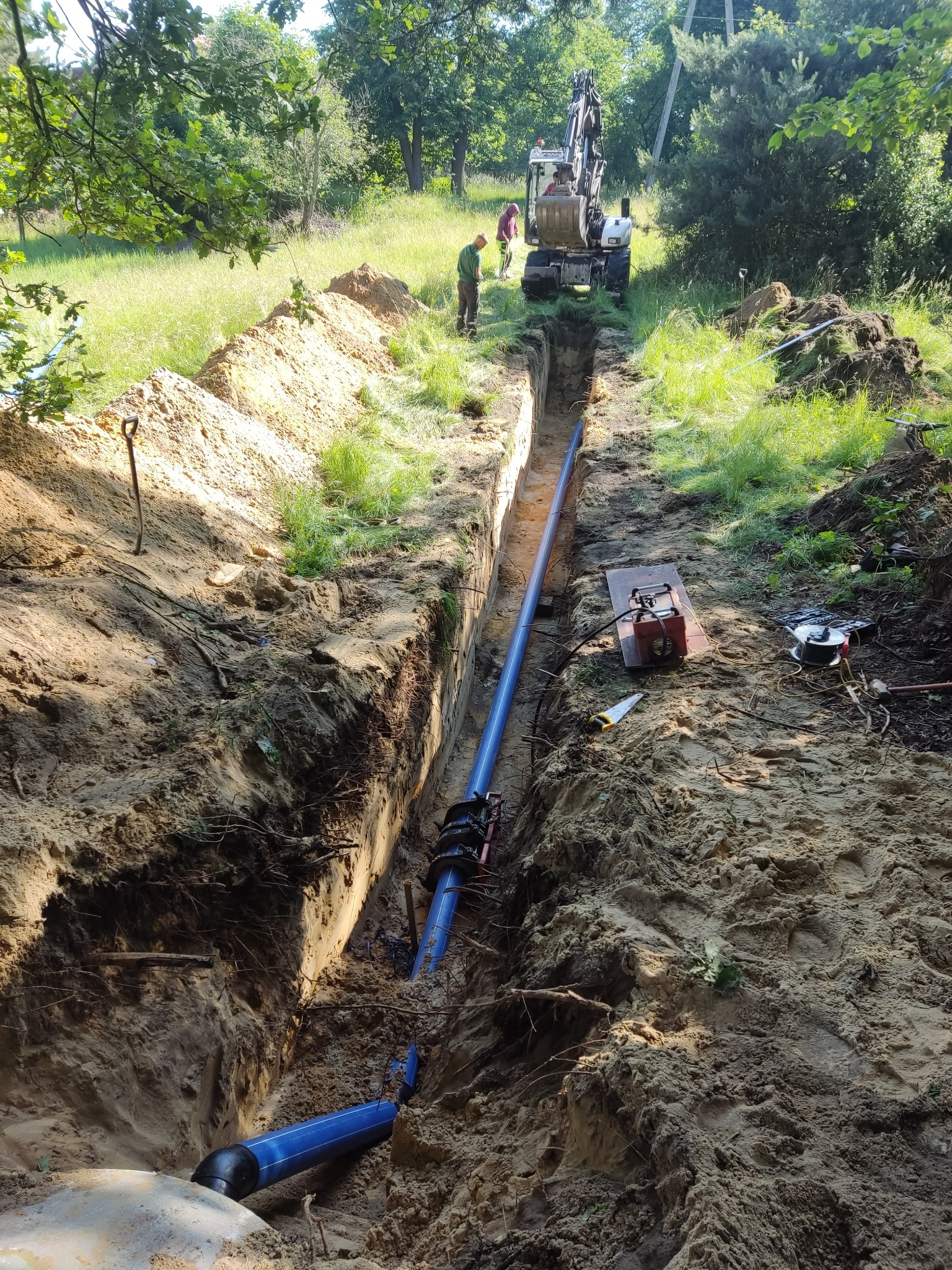 Głęboki wykop w ziemi z ułożonym niebieskim rurociągiem, widoczne łączenia rur, na górze wykopu leżą narzędzia, w tle pracująca koparka i osoby nadzorujące.