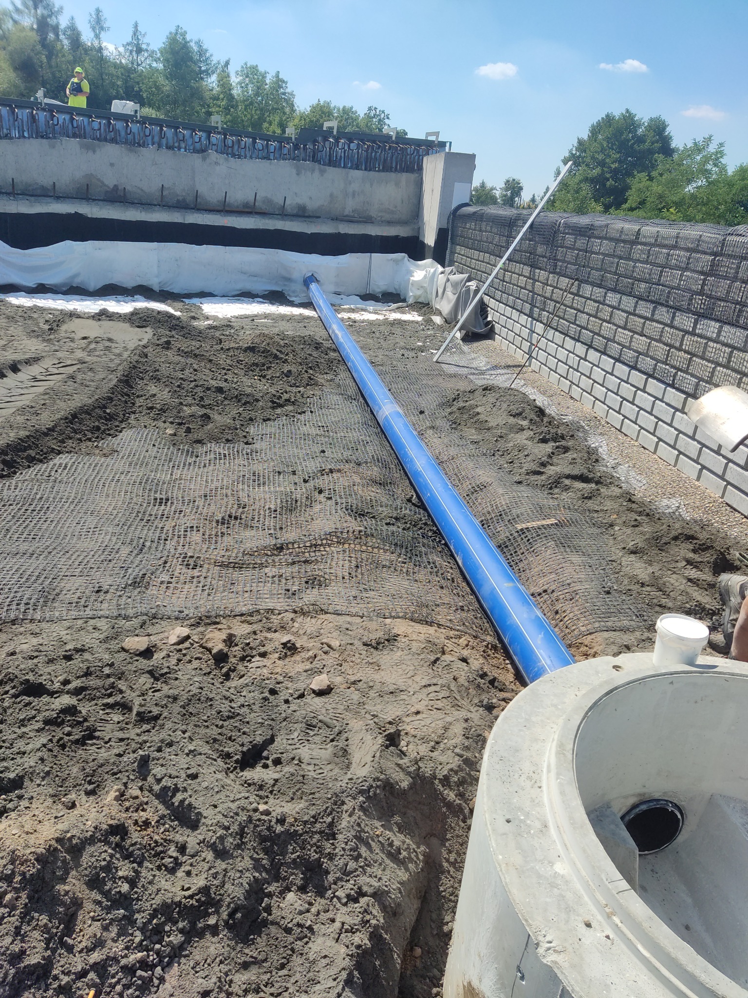 Niebieska rura leżąca na gruncie z siatką wzmacniającą, w tle betonowa konstrukcja z pracownikiem w żółtej kamizelce, z przodu betonowy element studni kanalizacyjnej.