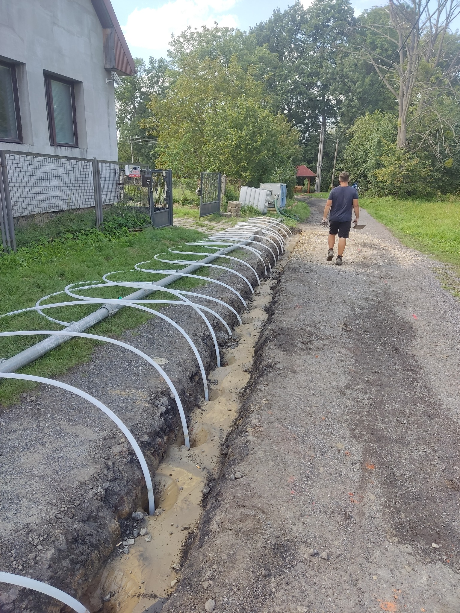 Rów z ułożonymi rurami w kształcie pętli, prawdopodobnie do pompy ciepła, z mężczyzną oddalającym się ścieżką w tle, widoczne budynki i zieleń.