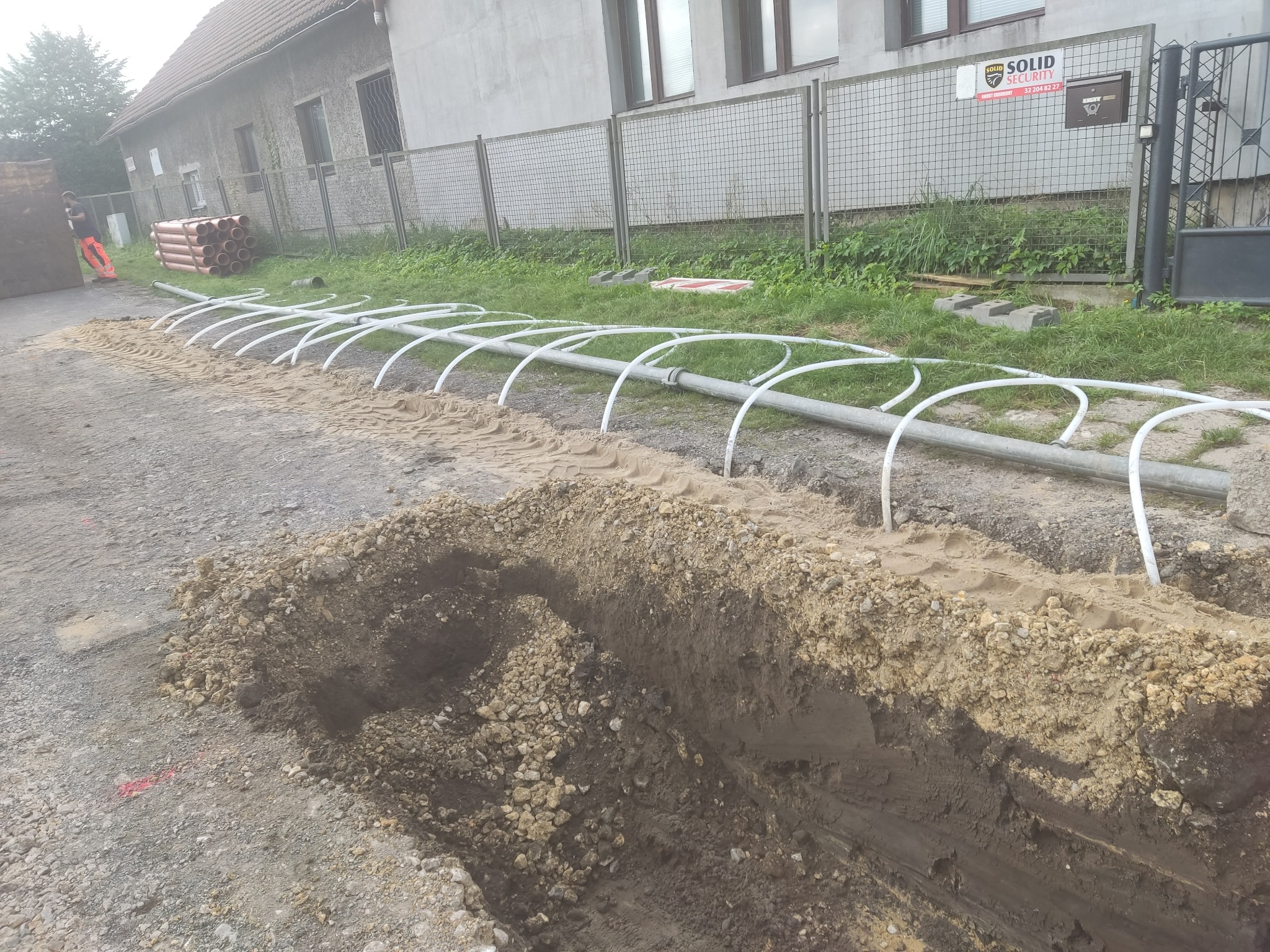 Wykopany rów z widocznymi rurami do podłączenia, w tle budynek i ogrodzenie z siatki.